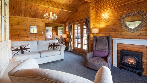 A cozy log cabin interior with a fireplace, a sofa with British flag cushions, a patterned armchair, and a chandelier.