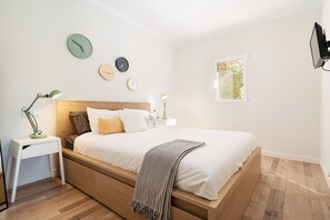 A bedroom with a wooden bed, white bedding, a bedside table with a lamp, and a window with a view of greenery.