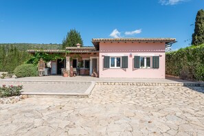 A pink building with a tiled roof, a stone-paved entrance, and a garden area with potted plants.