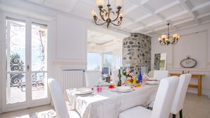 Un coin repas avec une table dressée pour quatre personnes, des chaises blanches, un lustre et un mur en pierre.