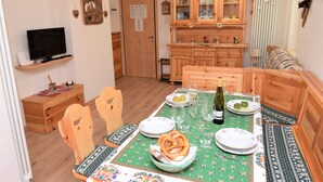 A dining area with a wooden table set for two, a television on a shelf, and a cabinet with glass doors.