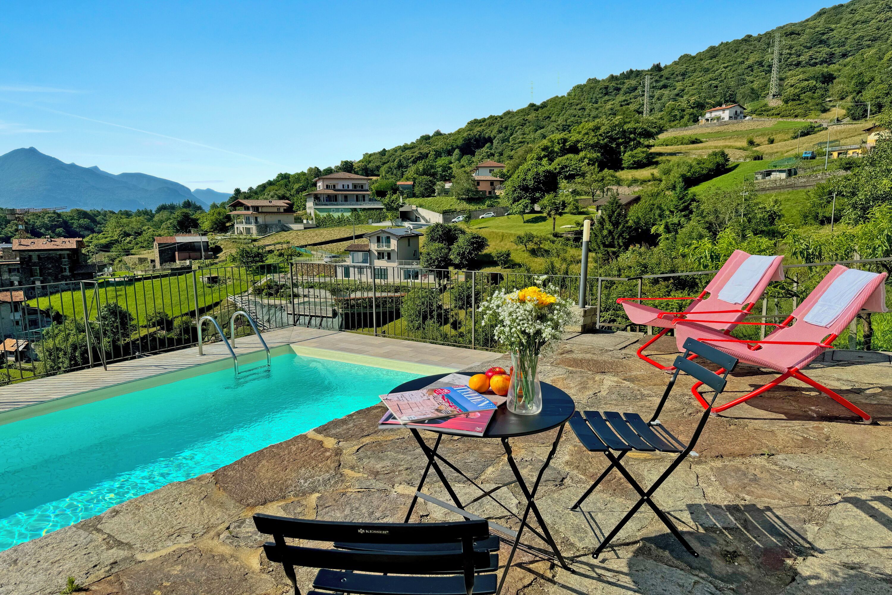 Zona de piscina con sillones de descanso y una mesa, rodeada de suelo de piedra y vistas a una ladera con casas.