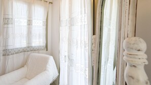 Una habitación con un sofá blanco, una puerta de madera con un pomo redondo y una ventana con cortinas translúcidas.