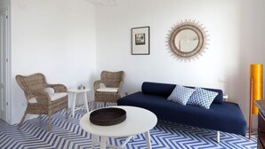 Une pièce avec un canapé bleu, deux fauteuils en osier, une petite table blanche et un miroir décoratif accroché au mur.
