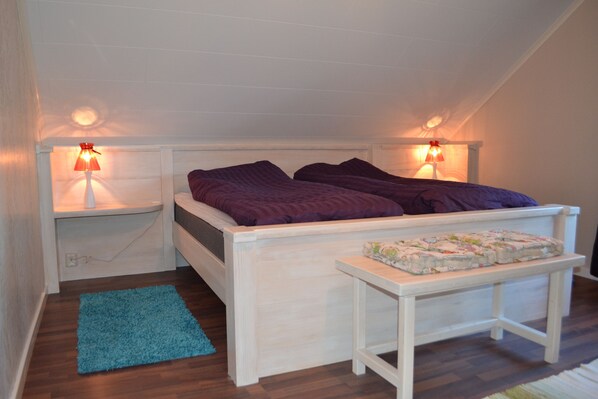 Une chambre avec un lit blanc, une literie violette, deux lampes de chevet, un tapis bleu et une petite table sur laquelle sont empilées des couches.