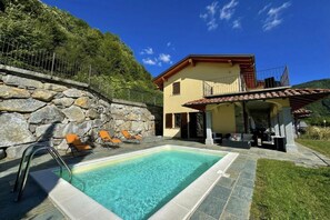 Un área de piscina con muros de piedra, un edificio amarillo con balcón y mobiliario exterior.