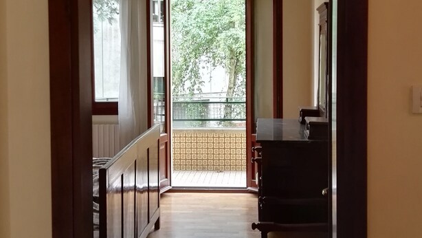 A hallway with a wooden floor, a ceiling-mounted air conditioner, a view of a balcony with greenery outside, and a dark-colored cabinet against the wall. - VENICE LIDO, SEA AND CINEMA<br>...  (Venezia)