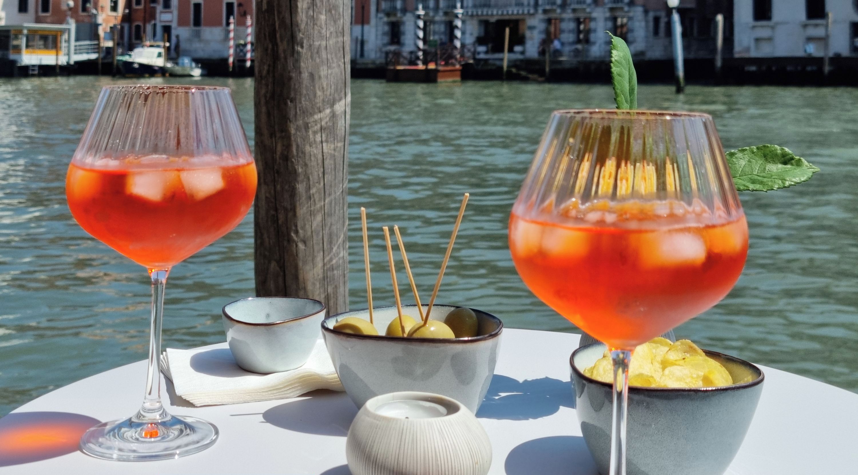 A table with two glasses of red cocktail, a bowl of olives, and a small candle. The background features a canal with boats and buildings.