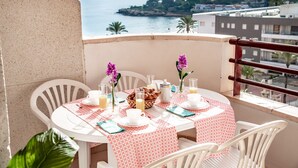 Un tavolo da balcone apparecchiato con prodotti per la colazione, sedie bianche e vista sul mare e sugli edifici.