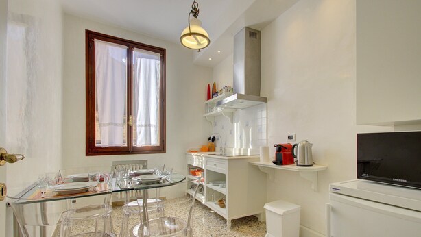 Uma cozinha moderna com uma mesa de jantar de vidro, cadeiras transparentes, forno embutido, micro-ondas e televisão.