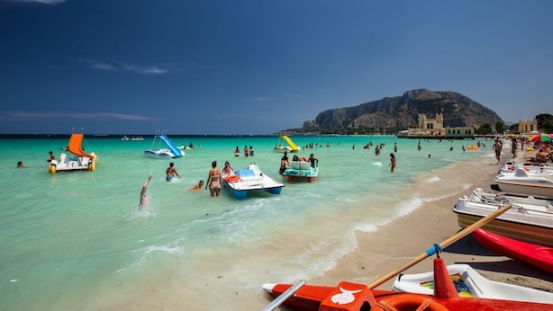 Una playa con agua turquesa cristalina, gente nadando y tomando el sol, y varios barcos.