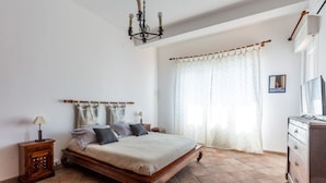 Un dormitorio con cama de madera, una mesita de noche, cómoda, candelabro y ventana con cortinas.