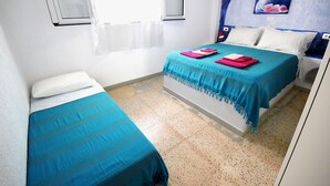 Ein Zimmer mit zwei Betten, eines größer, eines kleiner, beide mit blauer Bettwäsche. Ein Fenster mit weißem Vorhang, ein Wandbild und ein gefliester Boden sind ebenfalls vorhanden.