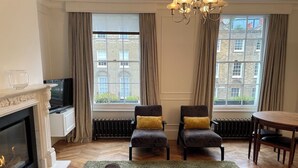 A living room with a fireplace, two armchairs, a sofa, a dining table, and a view of buildings through large windows. - Stylish in Angel, by Veeve (London)