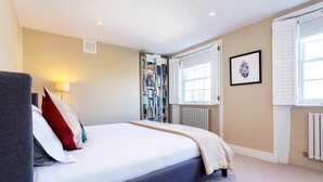 A bedroom with a bed, bedside lamp, bookshelf, and a window with shutters. - Stylish in Angel, by Veeve (London)