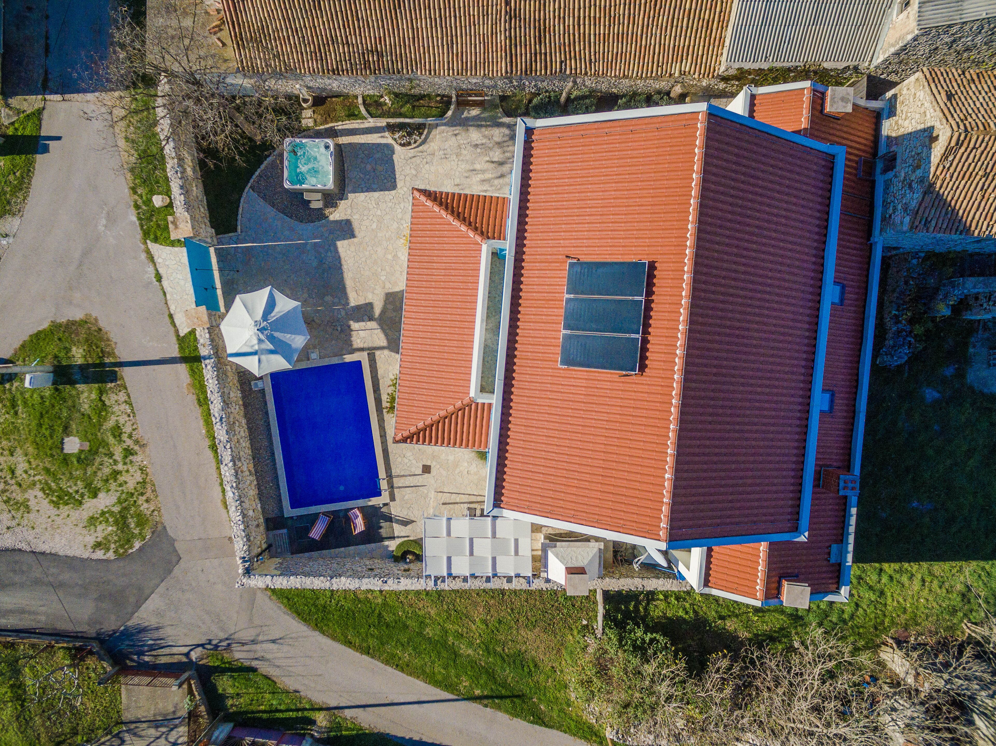 Vista aérea de uma casa com telhado vermelho, piscina azul e área de pátio com hidromassagem e guarda-sol.