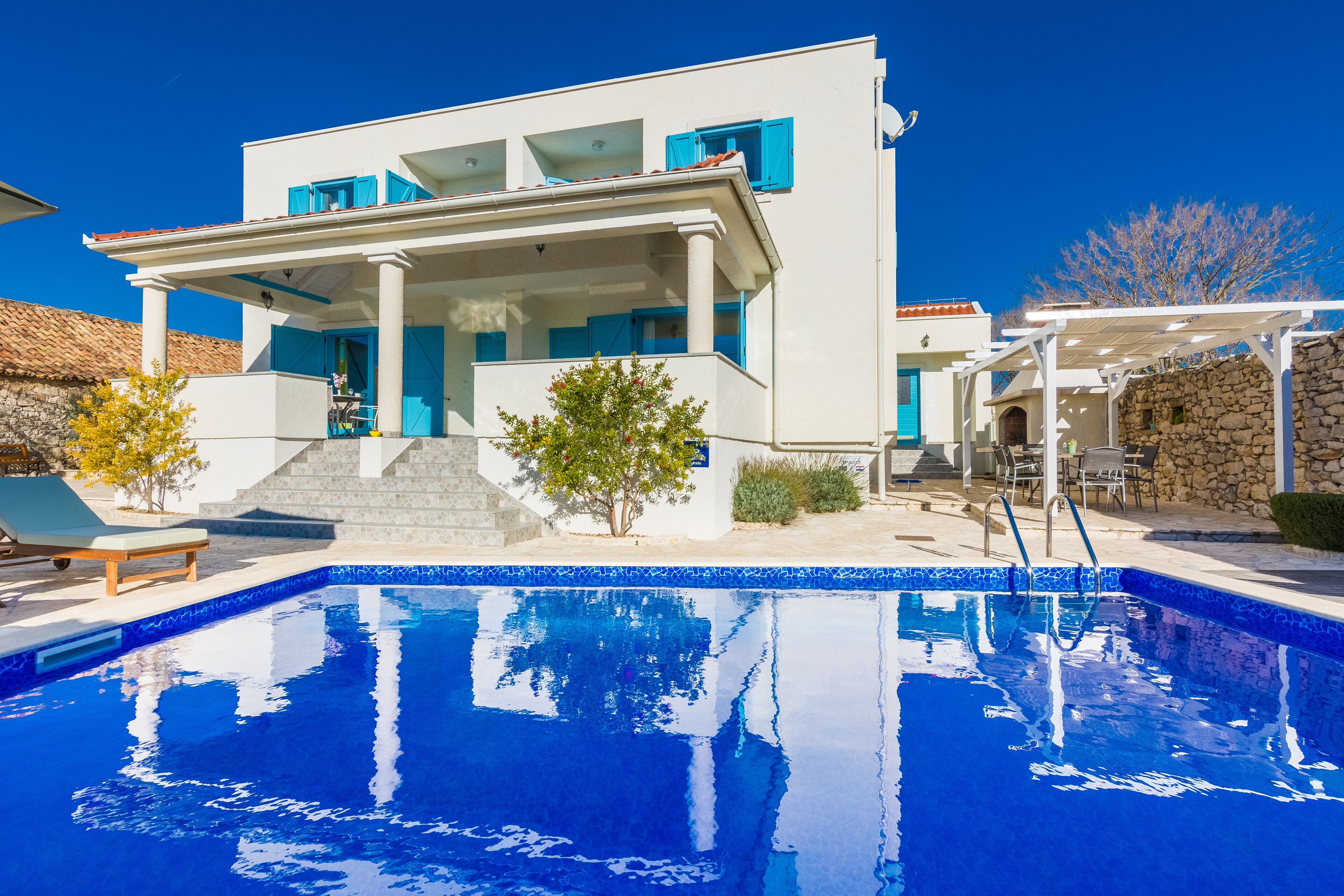 A modern villa with a swimming pool, a covered outdoor seating area, and a stone wall.