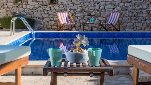 A poolside area with a stone wall, two lounge chairs, a small table with a plant, and two teal cups.
