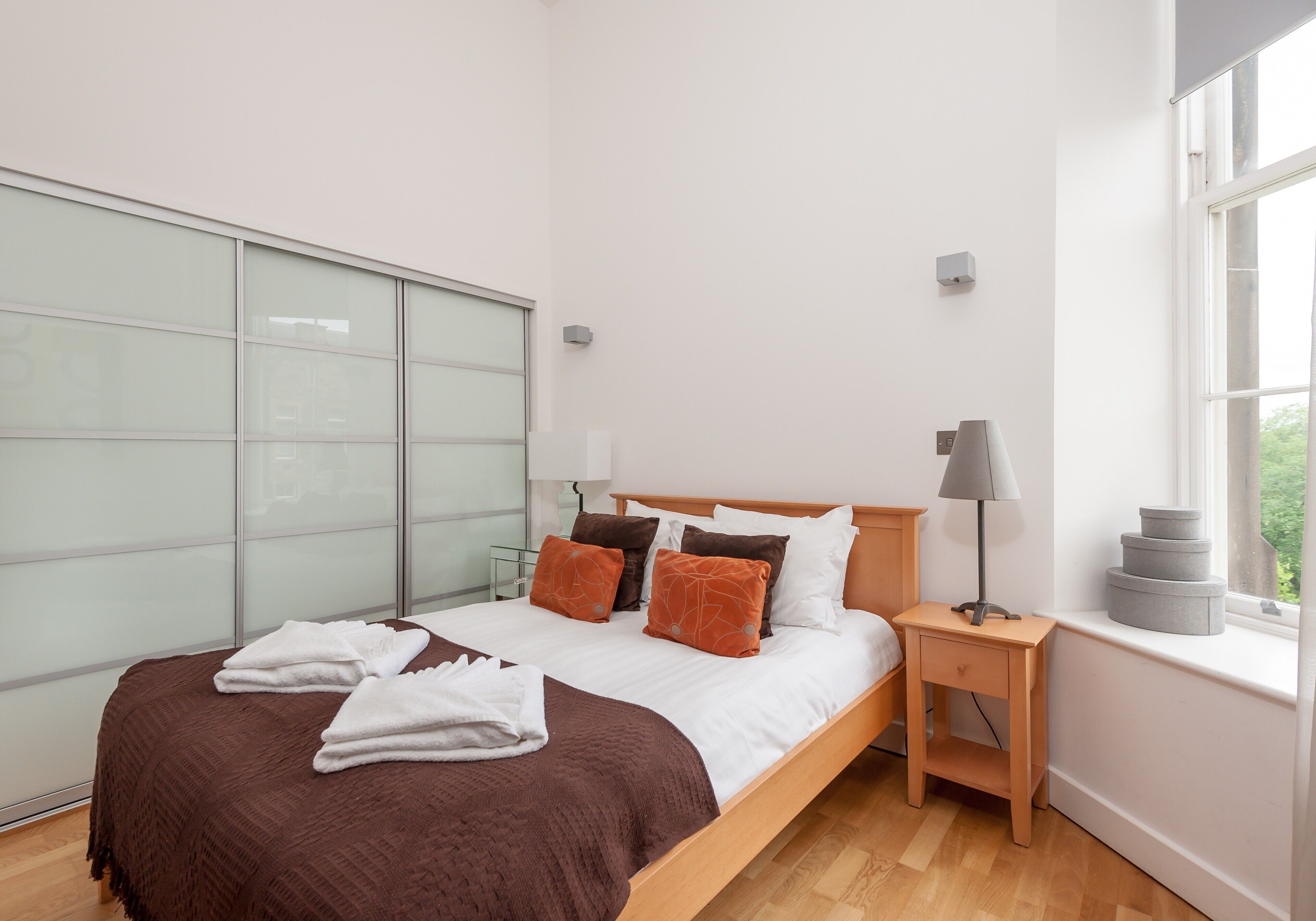 A modern bedroom with a large bed, a bedside table with a lamp, and a window with a view of greenery.