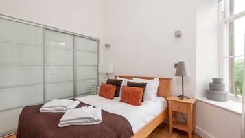A modern bedroom with a large bed, a bedside table with a lamp, and a window with a view of greenery.