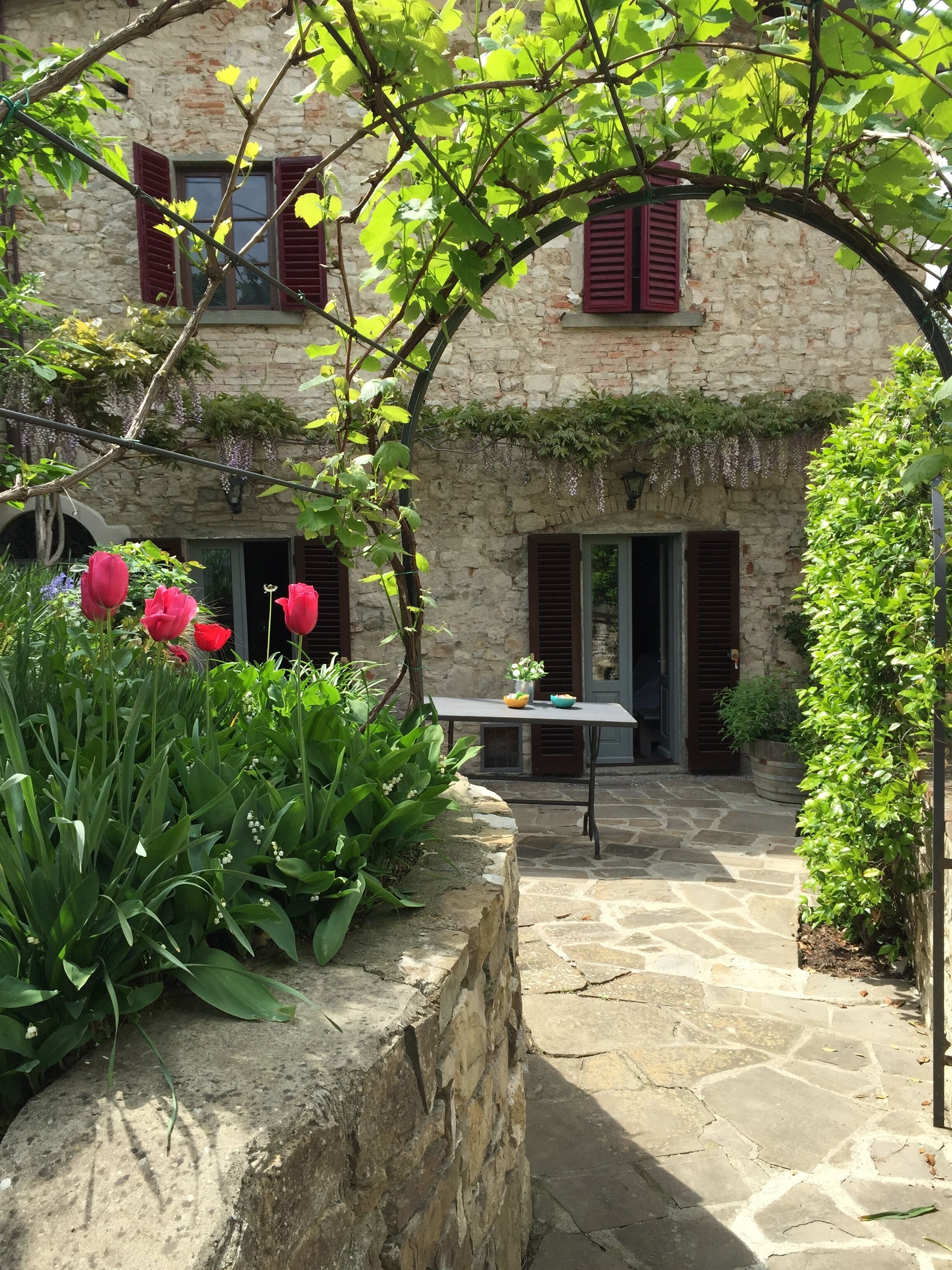 Ein Steinhaus mit Garten, einem Tisch mit Vase und Schale sowie roten Fensterläden.