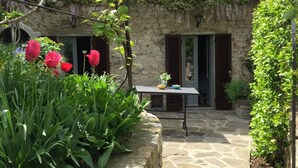 Un edificio de piedra con jardín, una mesa con un jarrón y un cuenco, y contraventanas rojas.