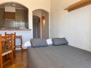 Un dormitorio con cama, una silla de madera, una zona de cocina con salpicadero de azulejos y un pasillo.