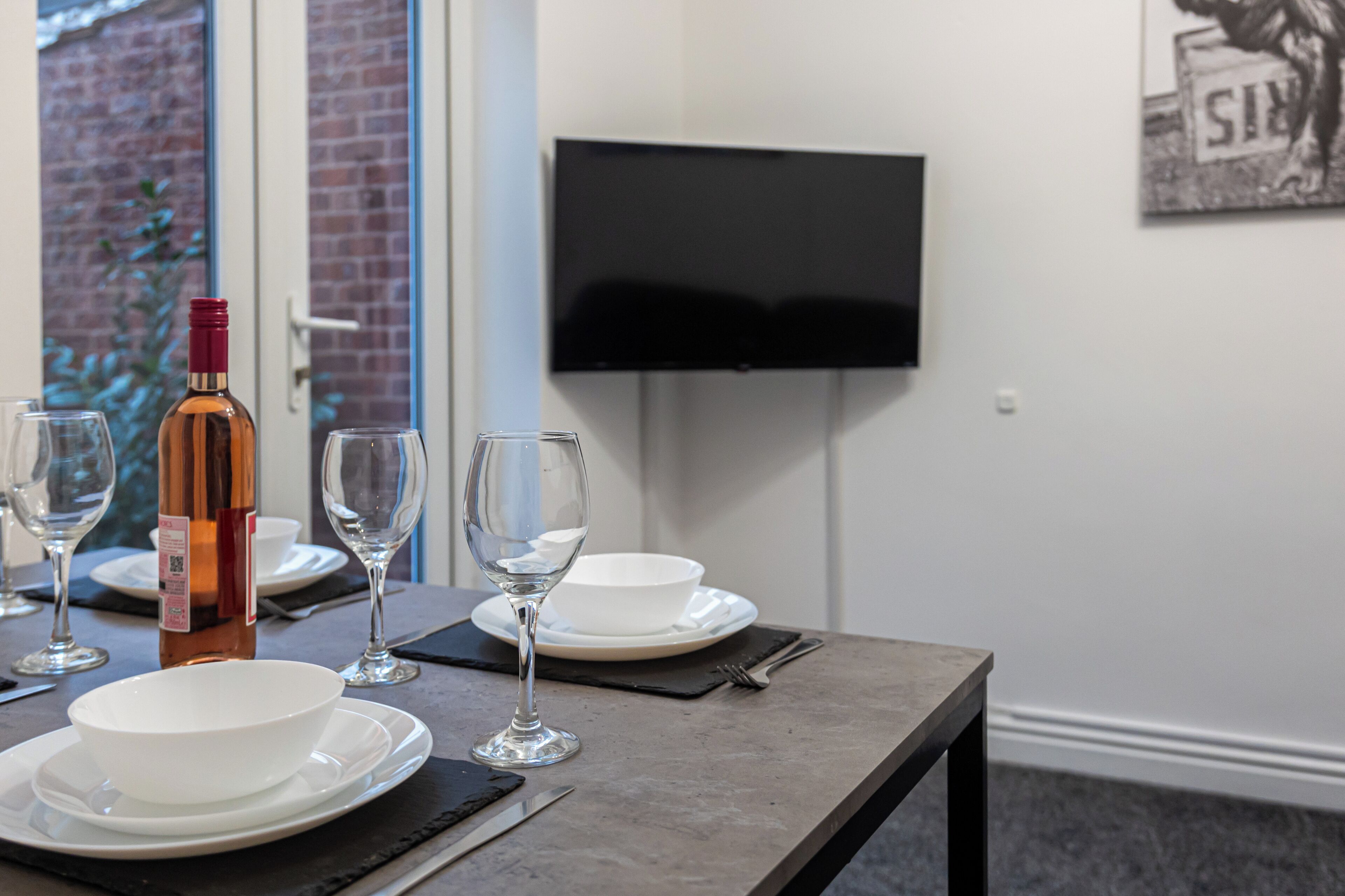 A dining table set with white plates, bowls, and wine glasses, a bottle of wine, and cutlery. A flat-screen TV is mounted on the wall above the table.
