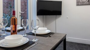 Mesa de jantar posta com pratos brancos, tigelas e copos de vinho, uma garrafa de vinho e talheres. Uma TV de tela plana está fixada na parede acima da mesa.