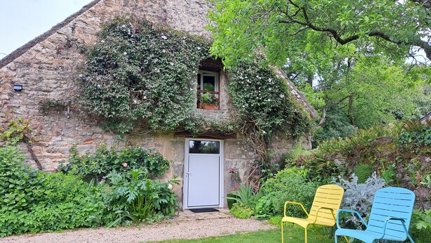 A stone house with a door and a small window, surrounded by lush greenery and two colorful chairs.