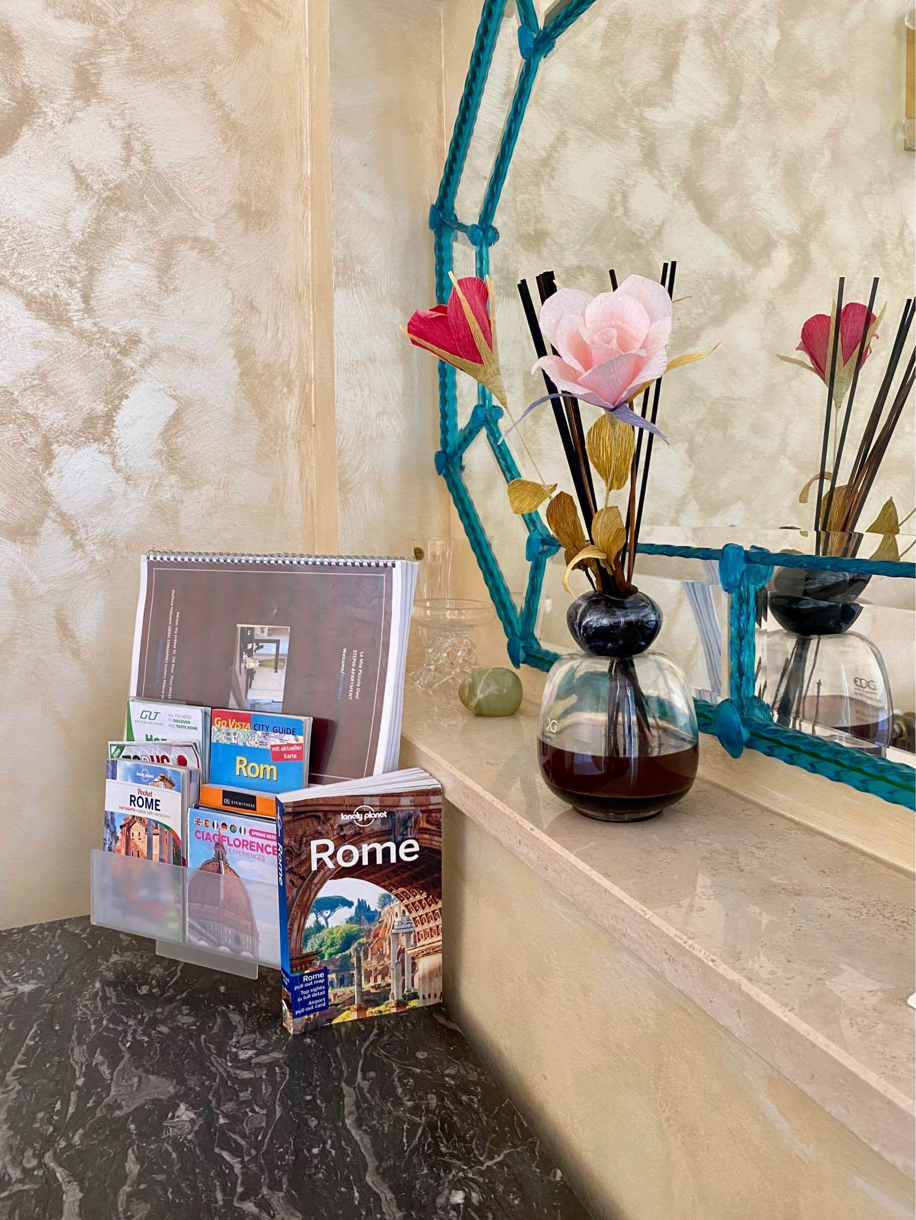 Una estantería con guías de viaje, un florero con flores y un espejo decorativo.