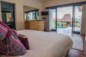 A bedroom with a large bed, a dresser, a TV, and a sliding glass door leading to a pool area. - Canggu Villa & Cooking Retreat (Tibubeneng)