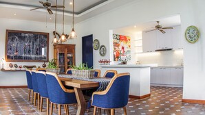 A dining area with a wooden table and blue upholstered chairs, a kitchen with white cabinets, and a ceiling fan. - Canggu Villa & Cooking Retreat (Tibubeneng)