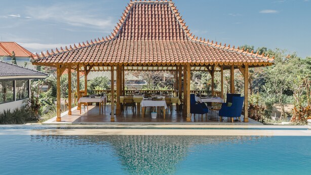 A covered outdoor dining area with a tiled roof, tables, and chairs, adjacent to a swimming pool. - Canggu Villa & Cooking Retreat (Tibubeneng)