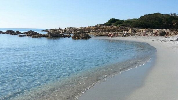 A beach with clear shallow waters, a sandy shore, and rocky outcrops in the background. - Villa Paradiso, in the beach by foot, big garden,  Wifi, all inclusive (Cala Liberotto)
