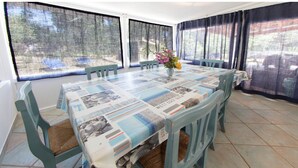 A dining area with a long table covered with a newspaper print tablecloth, surrounded by green chairs with woven seats. - Villa Paradiso, in the beach by foot, big garden,  Wifi, all inclusive (Cala Liberotto)