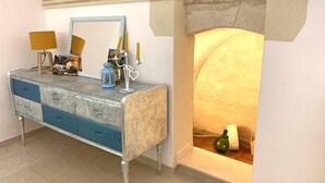 Habitació amb una llar de pedra, una petarrera blau i blanca amb calaixos, un mirall, una làmpada groga i un fanal blau.