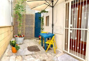 A small outdoor patio with a blue table and yellow chairs, potted plants, and a white door with a metal gate. - Al Vicoletto -Typical apartment in the historic center located on the ground floor (Ginosa)