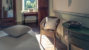 A bedroom with a bed, a wicker chair, a dresser, and a window with a view of trees. - Casale Amati La Marchesa, ancient and fascinating oil mill between sea and hills. (Ortonovo)