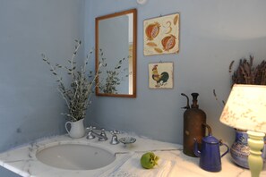 Kylpyhuone, jossa on pesuallas, peili, koristeellinen seinätaideteos, ruukkukasvi ja vanhanaikaistyylinen lamppu.