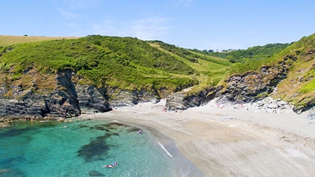 Une plage côtière aux eaux turquoise et limpides, bordée de sable fin, avec des collines verdoyantes en arrière-plan.