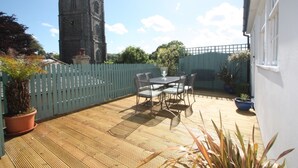 Une terrasse avec une table et des chaises, une plateforme en bois, une clôture blanche et, au loin, le clocher d’une église.