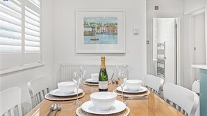 Une salle à manger avec une table en bois dressée pour six personnes, des chaises blanches et un tableau encadré au-dessus de la table.