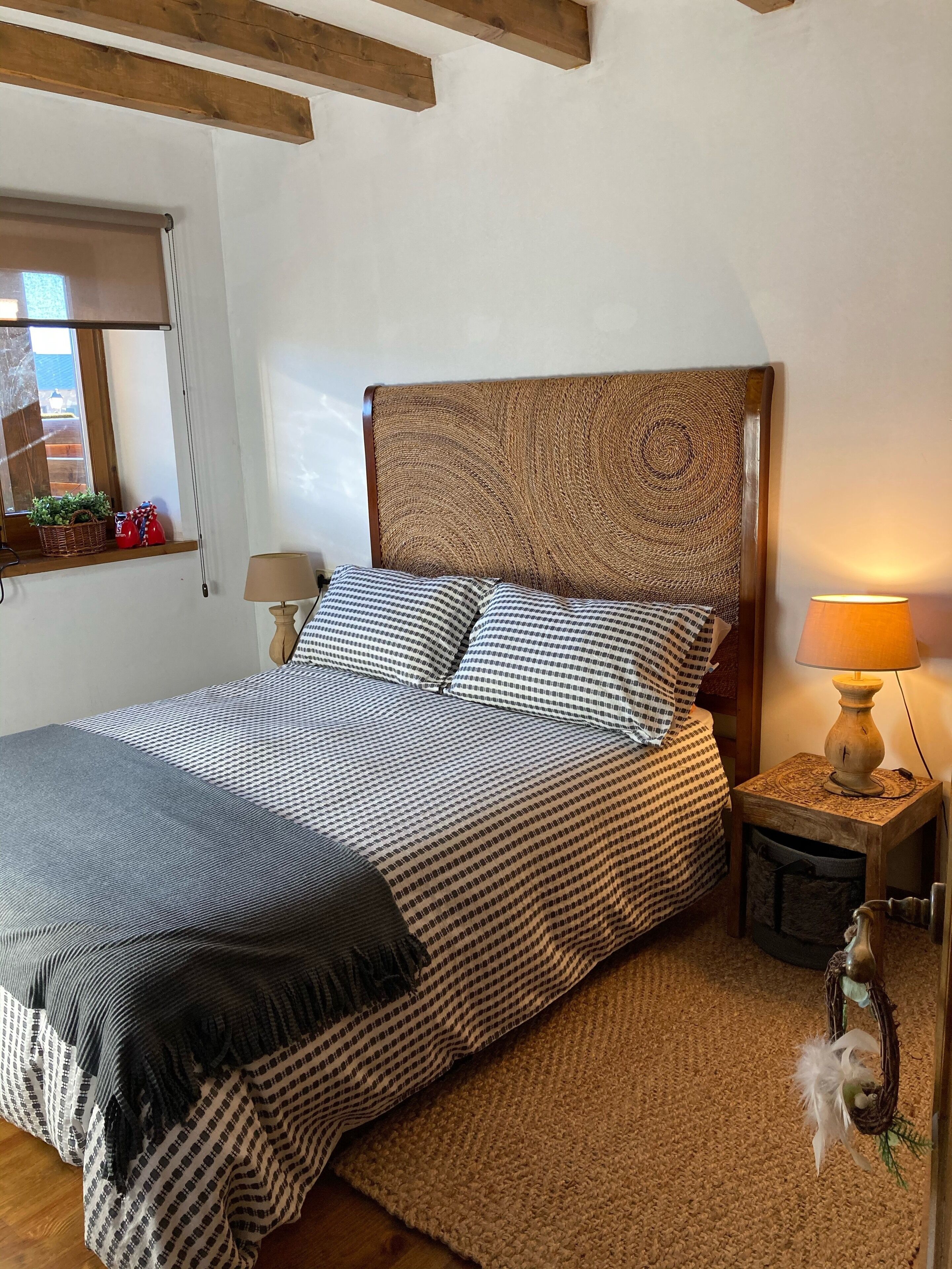Een slaapkamer met een bed, houten hoofdbord, nachtkastje met een lamp en een raam met uitzicht.
