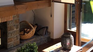 Una sala de estar con una televisión sobre un estante de madera, una chimenea con un cesto de leña, una planta en maceta y una ventana con vistas al exterior.