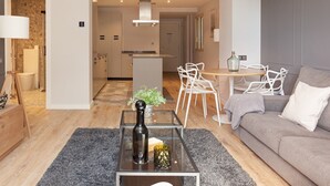 A modern living room with a glass coffee table, a grey sofa, and a dining area with white chairs.