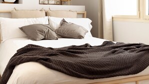 A neatly made bed with a dark blanket, a wooden headboard, and a bedside lamp.
