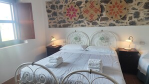 Un lit avec un cadre en métal, deux tables de chevet avec des lampes, un mur en pierre orné d’assiettes décoratives et une fenêtre avec des rideaux.