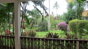 A covered outdoor area with a view of a lush garden, including various plants and a small pink flower bush.