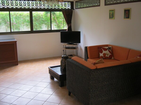 A living room with a wicker sofa, a television on a stand, a coffee table, and a view of greenery through the window.